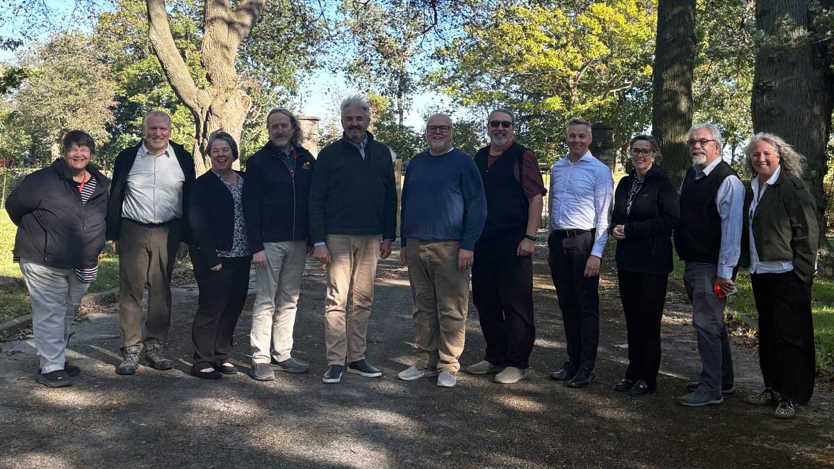 Eleven people stand together outdoors beneath large trees, smiling for a group photo on a sunny day.